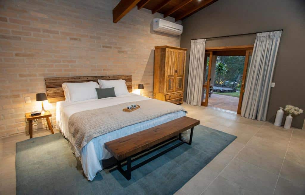 Quarto do Canto do Irere – Boutique Hotel com cama de casal ao centro com duas cômodas de madeira com luminária ao lado, no pé da cama tem um banco de madeira e do lado esquerdo do quarto portas amplas com cortinas brancas com acesso à varanda.