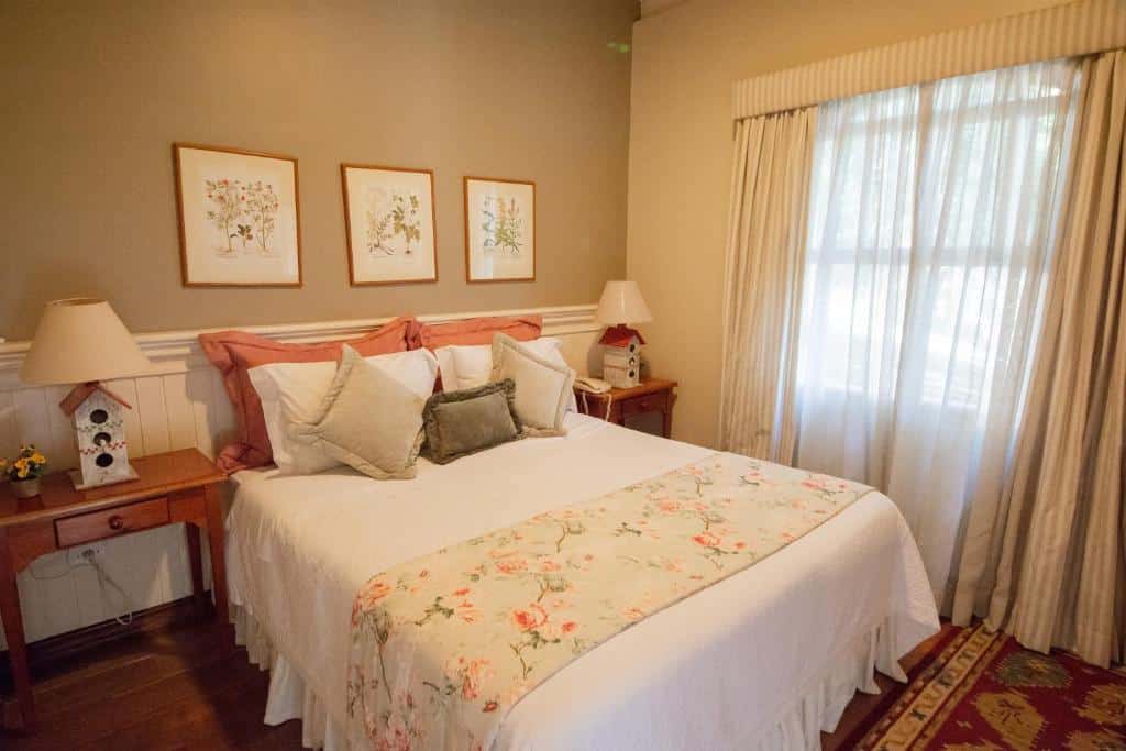 Quarto do Recanto Da Paz Hotel Fazenda com cama de casal no centro do quarto, duas cômodas de madeira ao lado da cama com luminária e do lado esquerdo janelas com cortinas brancas.
