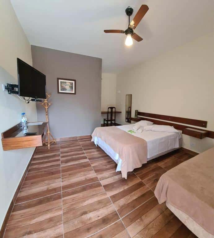Quarto do Estância Canaã com cama de casal do lado esquerdo, uma pequena cômoda de madeira ao meio, uma cama do lado direito, em frente a cama TV presa na parede e uma cômoda de madeira.