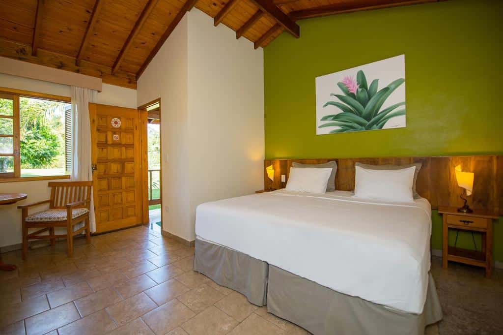 Quarto do Pousada Paiol com cama de casal do lado direito, com duas cômodas de madeira ao lado da cama com luminárias e do lado esquerdo uma cadeira de madeira perto da porta e da janela. 