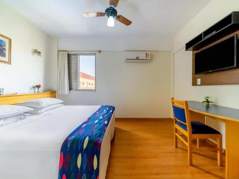 Quarto do Itapetinga Hotel durante o dia, com cama do lado esquerdo, e em frente a cama mesa de trabalho com cadeira e em cima TV presa na parede.