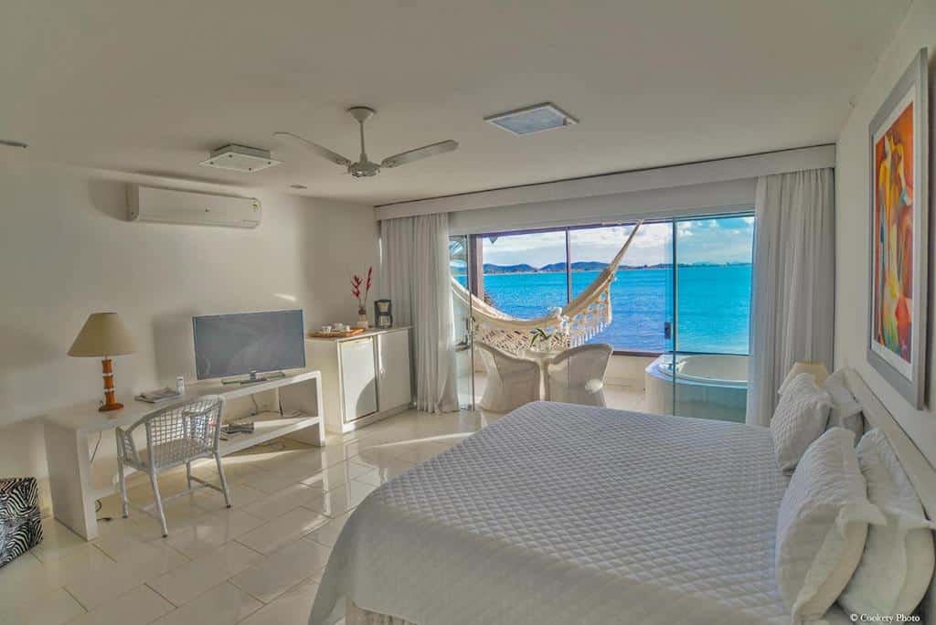 Quarto do Barracuda Eco Resort Búzios, com bastante espaço, há uma sacada com rede de descanso, uma banheira de hidromassagem e duas cadeiras, do lado de dentro, há uma cama de casal, uma televisão, um frigobar e um ventilador de teto