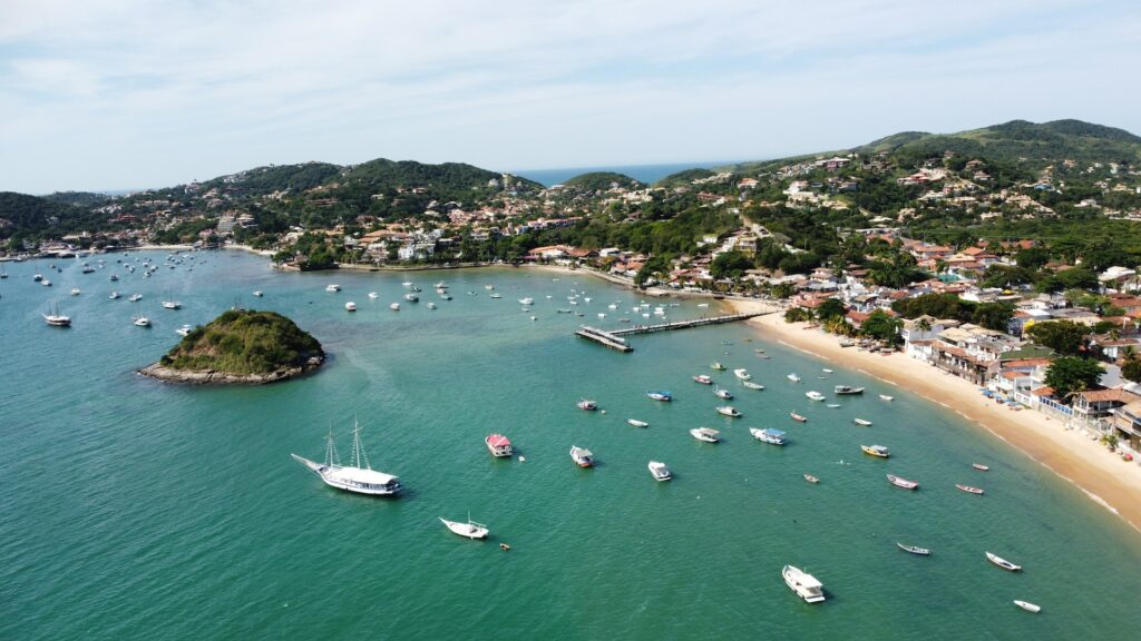 Vista aérea de uma praia com deck para embarcações, e há muitas no mar verde claro, e após a praia, há casas em meio as montanhas