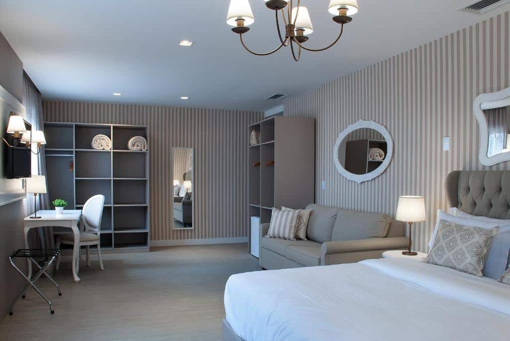 quarto do Bella Gramado by Gramado Parks com uma cama de casal à direita, um sofá ao lado da cama, um armário ao fundo, um espelho estreito na parede de trás e, à esquerda, uma pequena mesa branca com uma cadeira almofadada da mesma cor