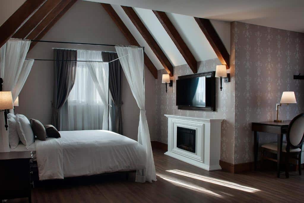 suíte do Buona Vitta Gramado Resort & Spa by Gramado Parks com uma cama de casal à esquerda, uma lareira branca em frente a cama, uma televisão de tela plana em cima da lareira, e uma mesa de madeira escura com uma cadeira almofadada à direita do quarto.