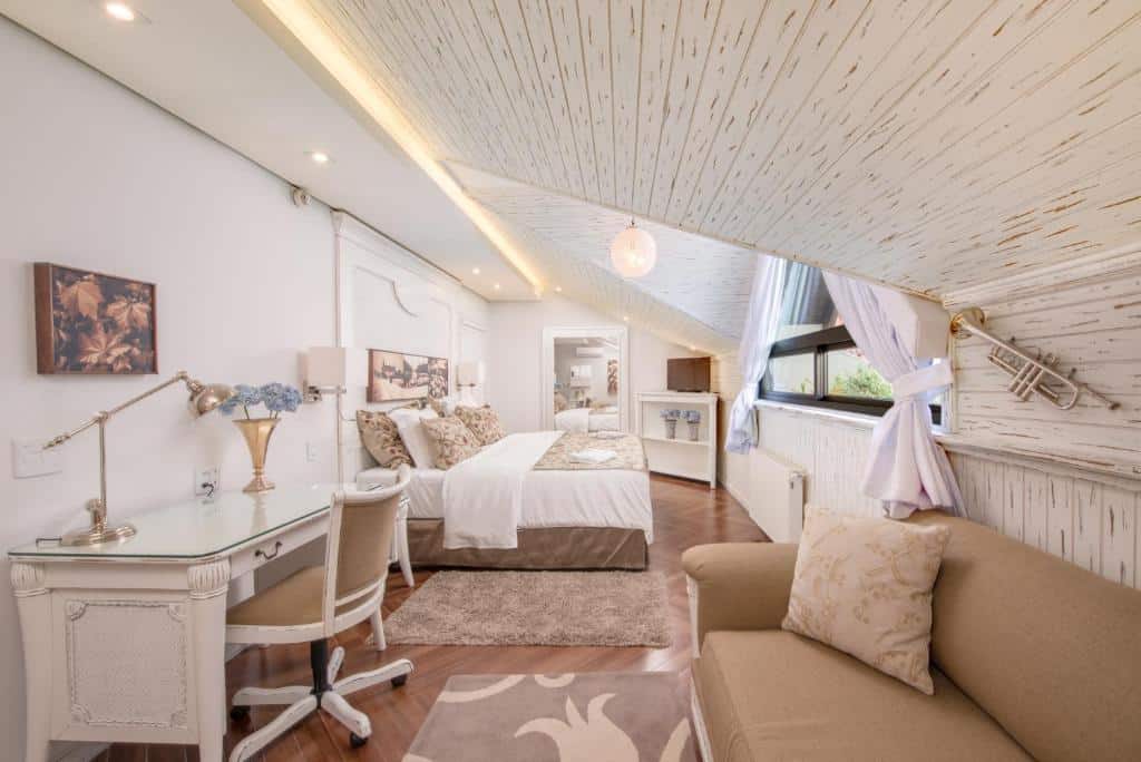 suíte do resort Wish Serrano em Gramado em tons branco e bege claro. Na frente, no canto direito, há um sofá e um trompete pendurado em cima, na parede. Ao fundo, há uma pequena cômoda branca de canto com uma televisão, ao seu lado já um espelho branco, grande e vertical. Em frente ao espelho, no lado esquerdo, está a cama de casal e, mais próximo do sofá, no lado esquerdo, há uma mesa branca de madeira com uma cadeira branca estofada e giratória na frente.