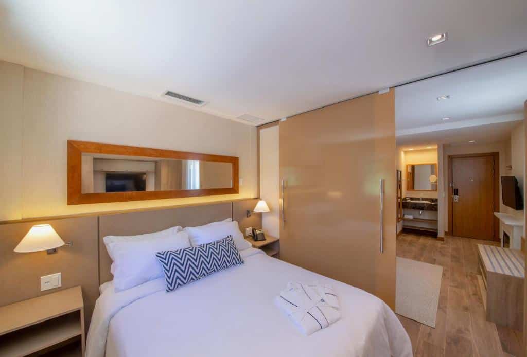 quarto do Wyndham Gramado Termas Resort & Spa com uma cama de casal à frente, uma porta de correr dividindo o quarto da sala com uma lareira branca e uma televisão posta em cima. Ao fundo é possível ver a porta de entrada e do lado esquerdo dela, uma pia com espelho que dá acesso ao banheiro.