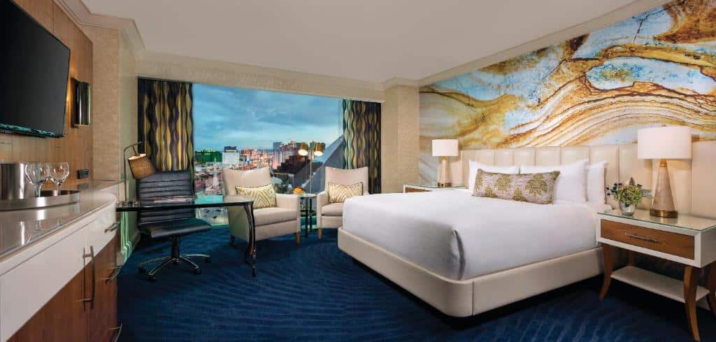 Quarto do Mandalay Bay com uma carpete azul-marinho, uma cama de casal com uma cabeceira bege, com mesinhas de cabeceira com abajur, há uma janela ampla com cortinas e duas poltronas de couro, do lado esquerdo há uma mesinha de escritório com uma cadeira, além de uma televisão e um móvel com gavetas