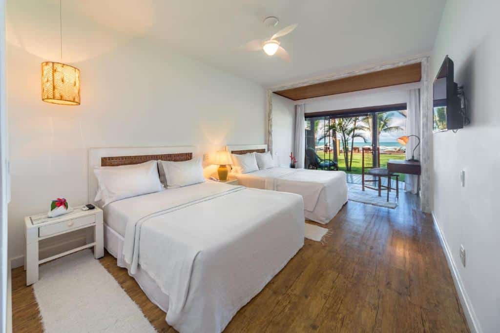 Quarto do Arraial D'ajuda Eco, um dos resorts para o Réveillon, com duas camas de casal, roupa de cama branca, piso de madeira, escrivaninha, TV e varanda com vista para as palmeiras e o oceano.