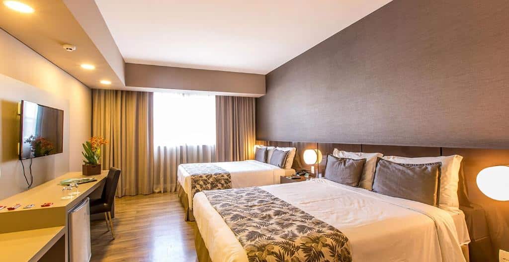 Quarto do Tauá Hotel & Convention Atibaia com duas camas de casal, escrivaninha, cadeira, TV de tela plana e janela com cortinas que permitem a entrada de luz natural.