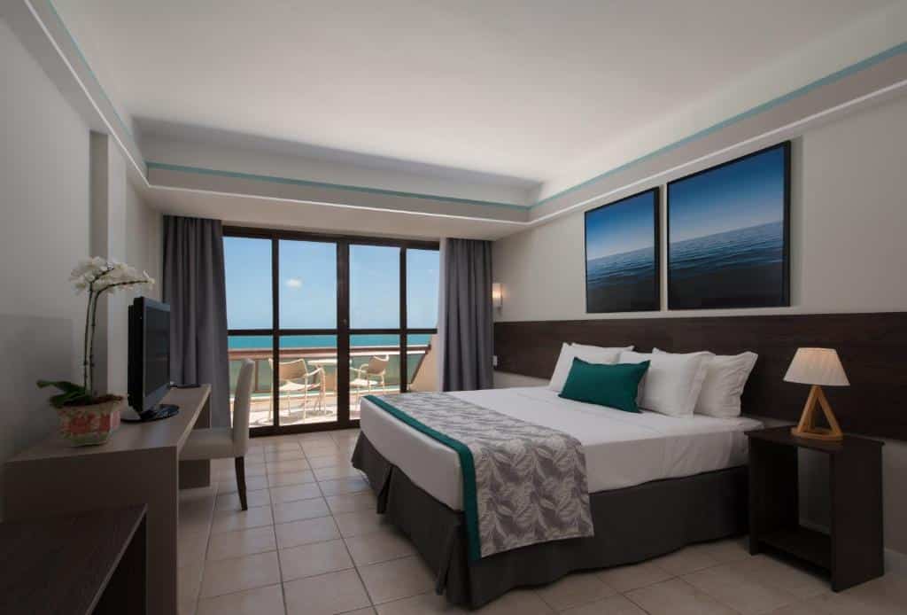 Quarto do Tauá Hotel & Convention Atibaia com uma cama grande, escrivaninha, TV, abajur e varanda com vista para o mar por meio de portas de vidro deslizantes.