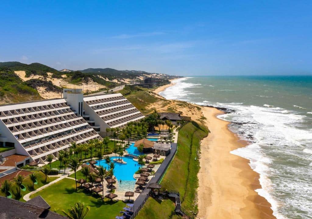 Amplo resort Tauá Hotel & Convention Atibaia à beira-mar com piscina, palmeiras e áreas de lounge ao lado de uma praia de areia e ondas do mar sob um céu claro. Um dos resorts para o Réveillon.