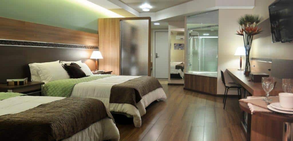 Quarto do Recanto Cataratas - Thermas, Resort e Convention com duas camas de casal, banheiro com paredes de vidro, piso de madeira, mesa de trabalho, TV e decoração moderna.