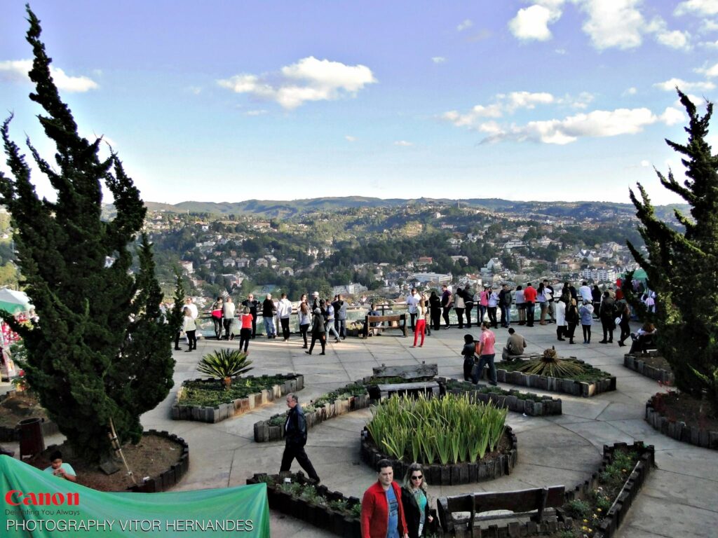 Espaço com árvores, pequenos jardins, bancos de madeira e um mirante com pessoas encostadas olhando a vista no Morro do Elefante em Campos do Jordão