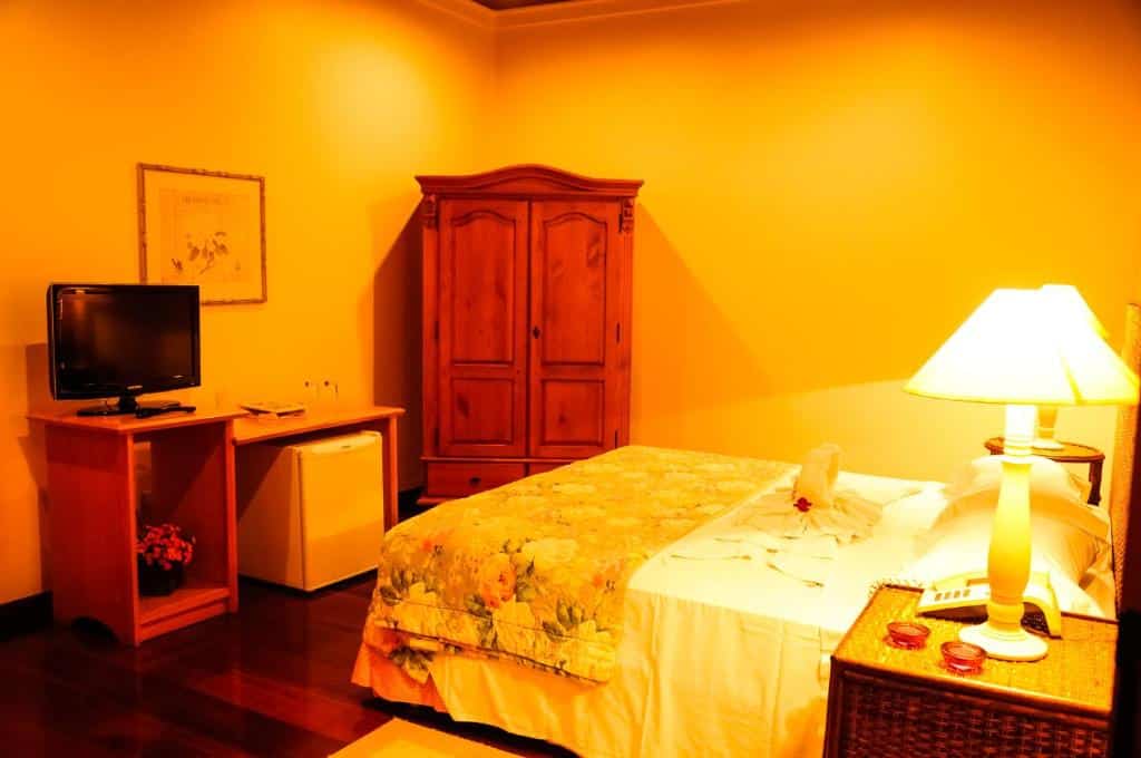 Quarto do Canto da Floresta Ecoresort com uma cama de casal, um pequeno armário de época de madeira, um frigobar, umaa pequena mesinha com uma televisão em cima, e nas laterias da cama há pequenas mesas de cabeceira com abajures em cima