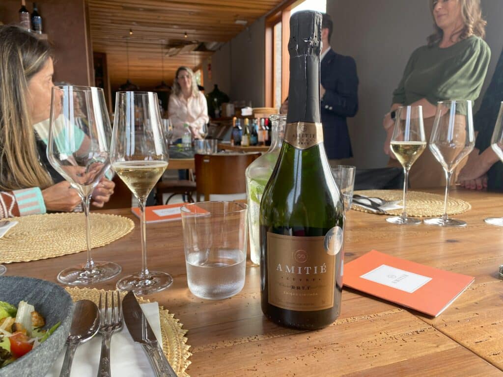 Garrafa de espumante Cuvée Brut da Amitié, uma das marcas de vinhos de Bento Gonçalves, apoiada sobre uma mesa de madeira, com taças ao redor
